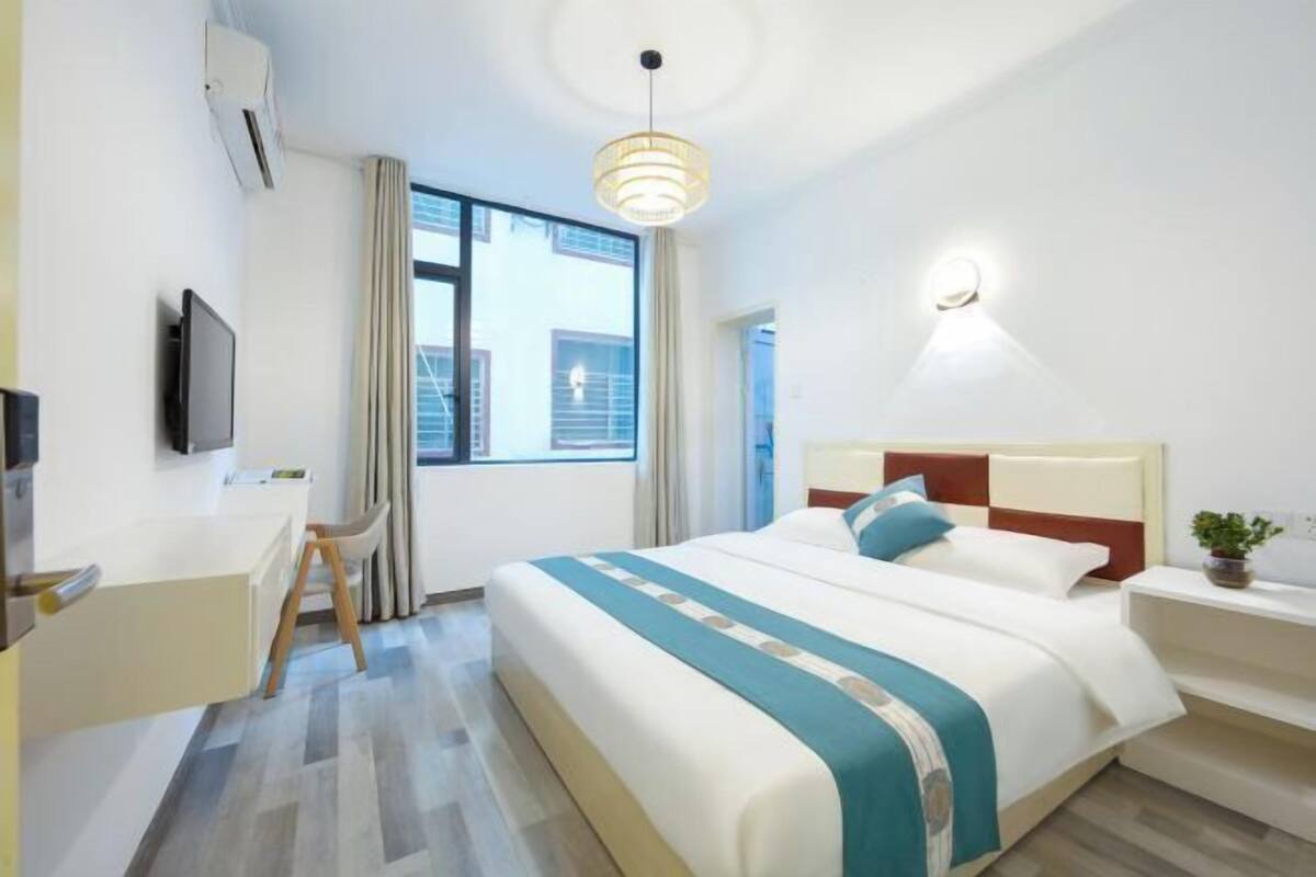 A hotel room with a bed, a desk, a chair, a TV, a window, and a wall-mounted light.