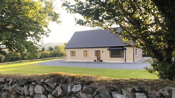 A house with a stone wall in the foreground, a well-maintained lawn, and a gravel driveway. - Charming 3-bedroom bungalow in Kenmare with WiFi access (Kenmare)