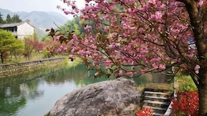 Un entorno tranquilo junto al río, con un edificio tradicional, un puente de piedra y una gran roca.