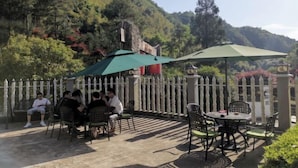 Un patio al aire libre con mesas y sillas, rodeado por una cerca blanca de piquete, con colinas verdes y exuberantes de fondo.