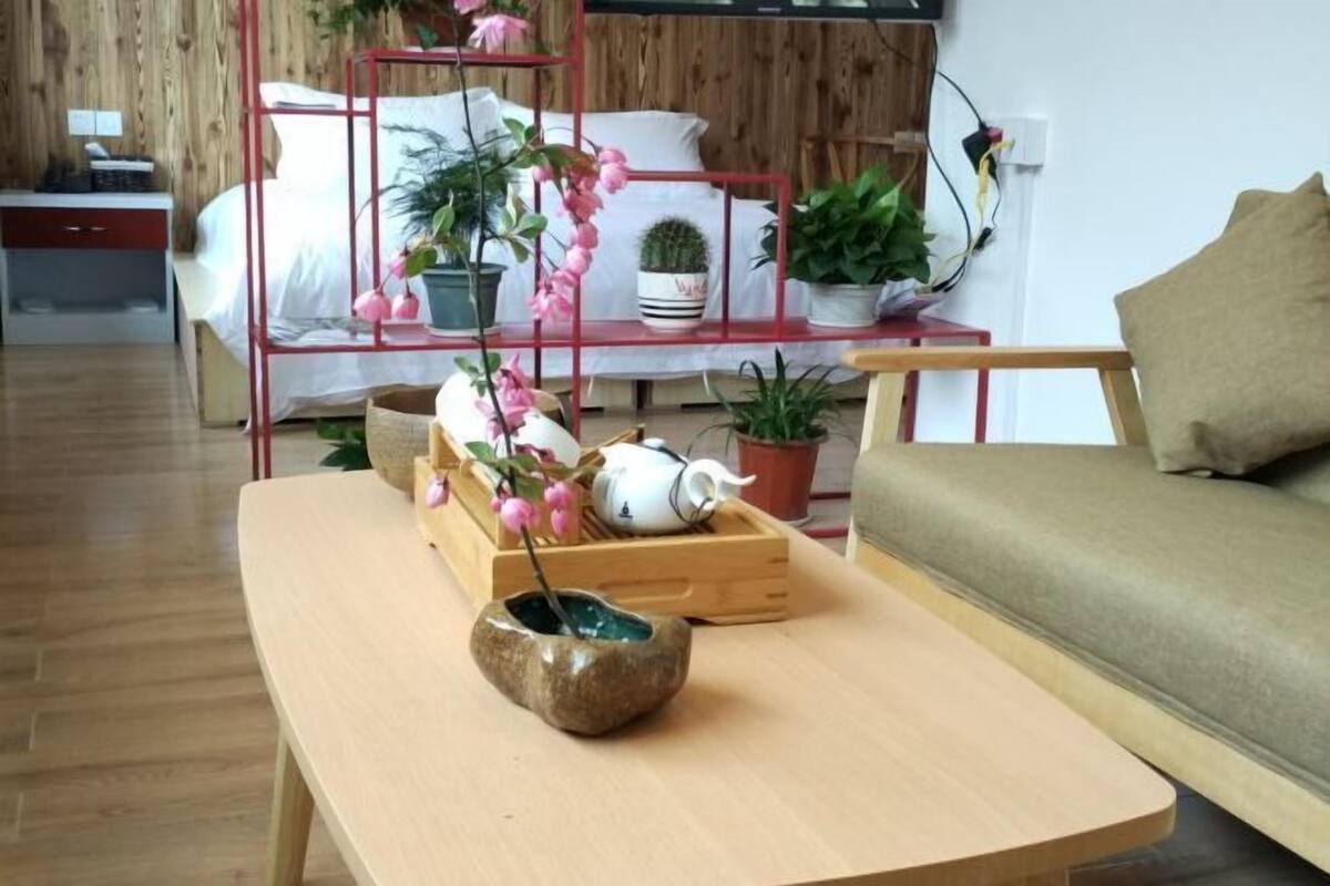 Une pièce avec un plancher en bois, un lit recouvert de draps blancs, une étagère en métal rouge ornée de plantes, une table en bois sur laquelle se trouvent une théière et un bol en pierre, et un canapé vert.