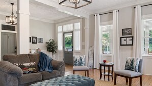 Amplia sala de estar con un sofá gris, un reposapiés azul y una pequeña mesa con una taza roja. Hay dos sillones con cojines decorativos, una planta en maceta y una puerta que da a otra habitación.