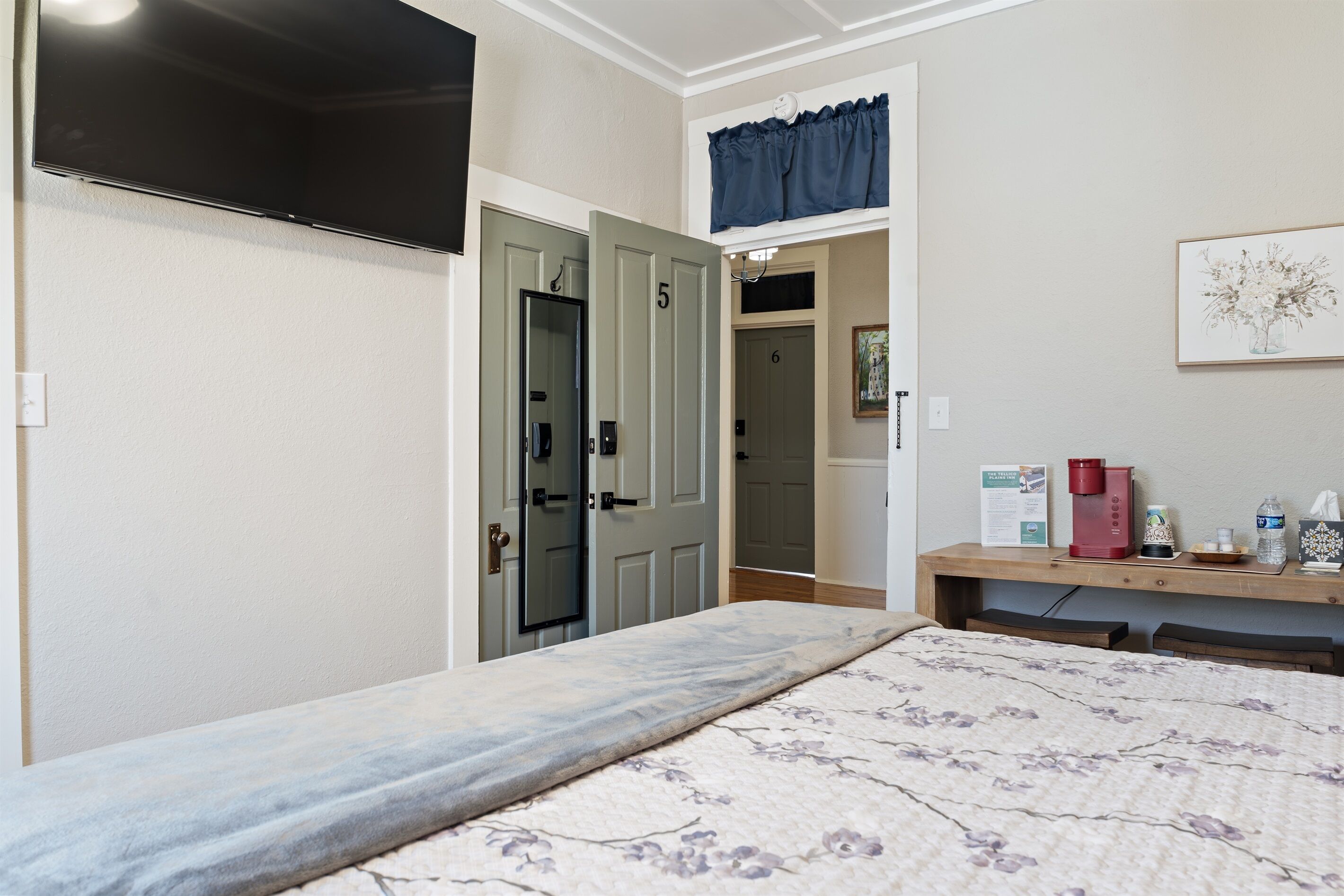 Habitación de hotel con cama, televisor, escritorio y cafetera, además de una puerta con números.