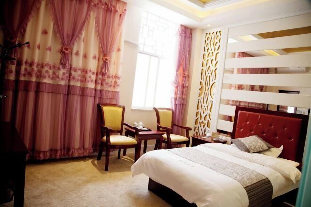 A hotel room with a bed, two chairs, a small table, a window with curtains, and a decorative wall panel.