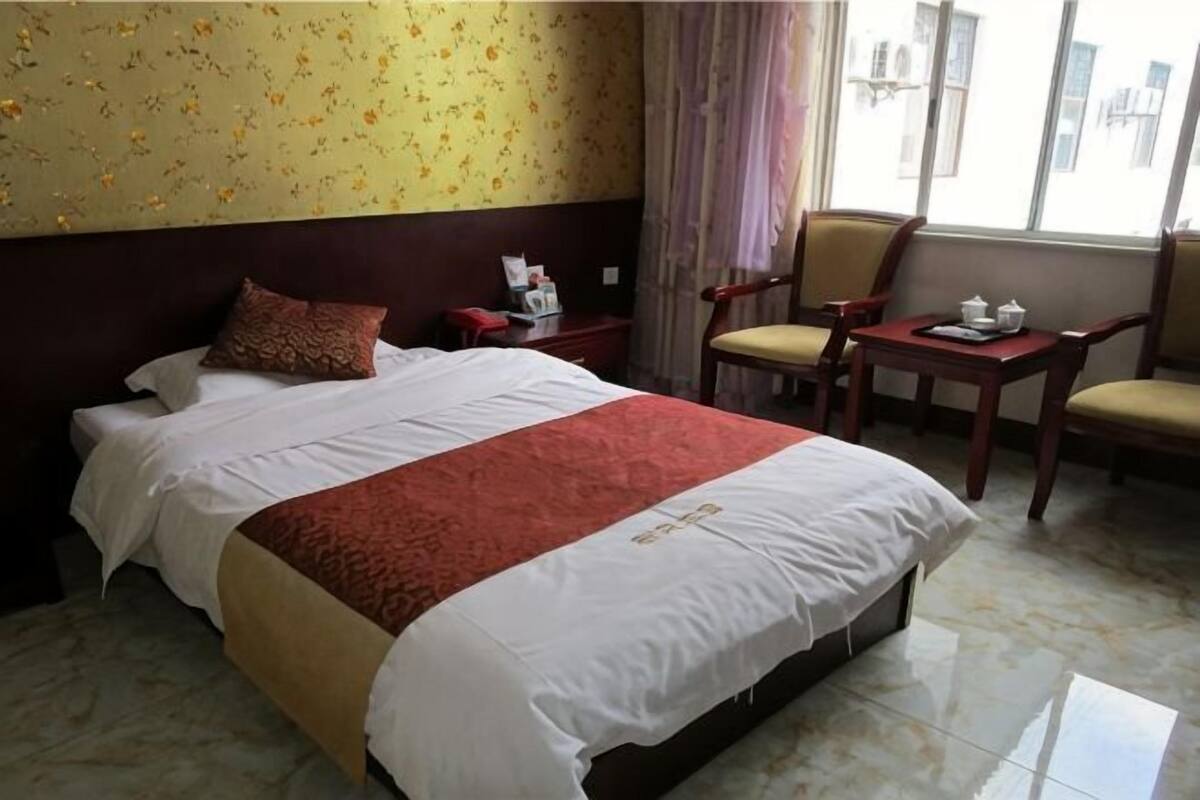 A hotel room with a large bed, a bedside table with a lamp, a chair, a small table with a teapot, and a window with curtains.