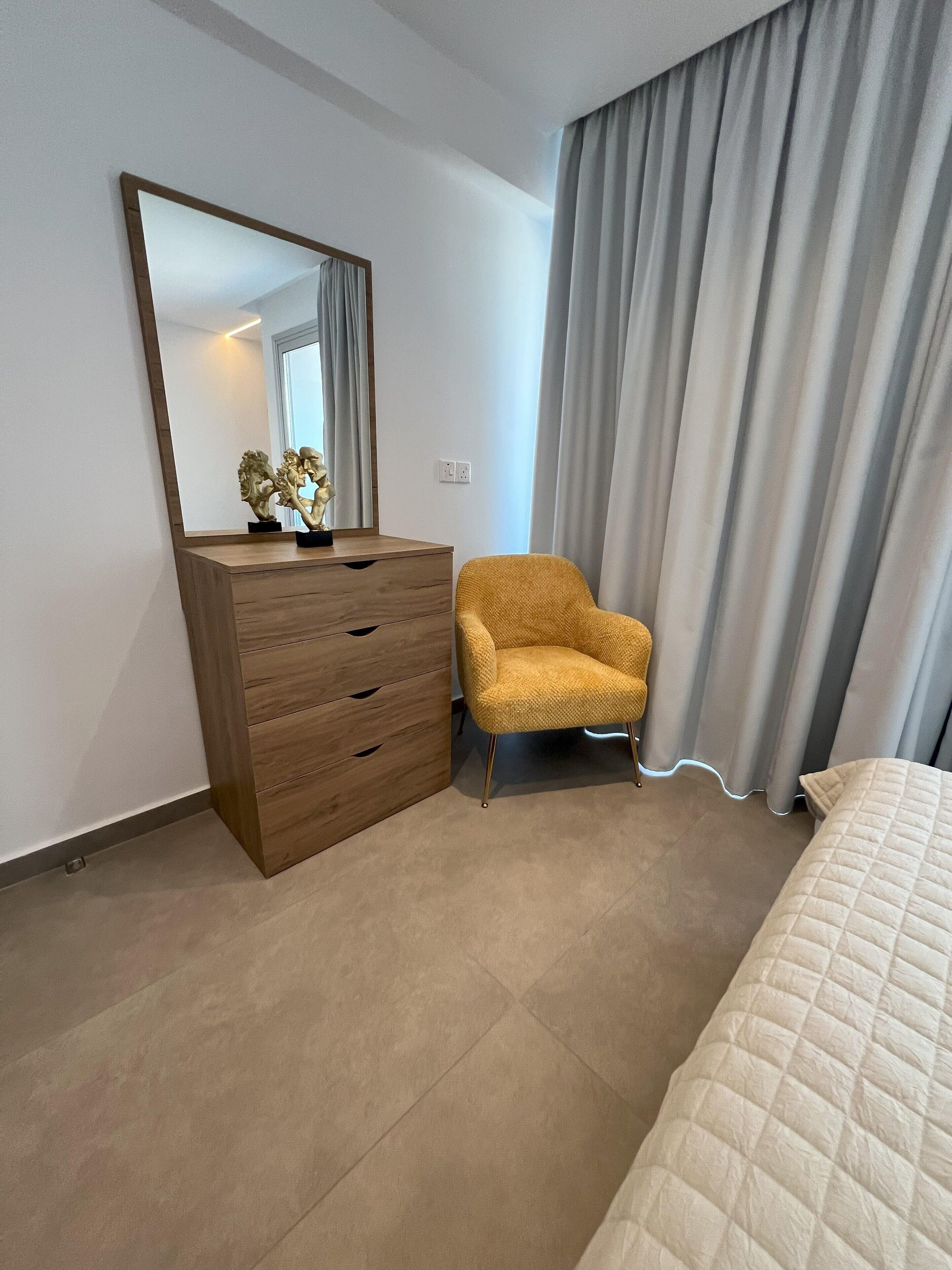 Une chambre à coucher avec une commode en bois, un fauteuil jaune, un lit et une grande fenêtre avec des rideaux.
