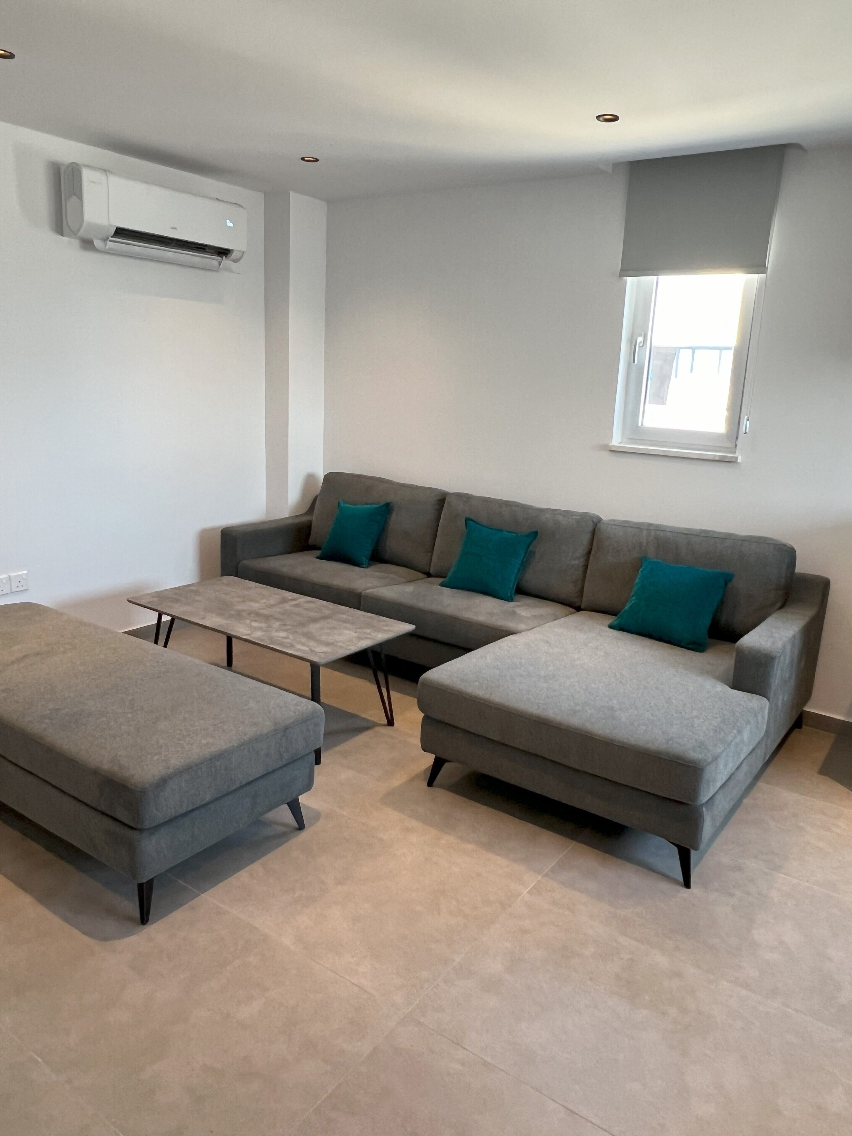 Ein modernes Wohnzimmer mit einem grauen Ecksofa, einem passenden Hocker und einem kleinen Couchtisch. Ein Fenster mit weißem Rollo, eine Klimaanlage an der Wand und ein Fliesenboden sind ebenfalls vorhanden.