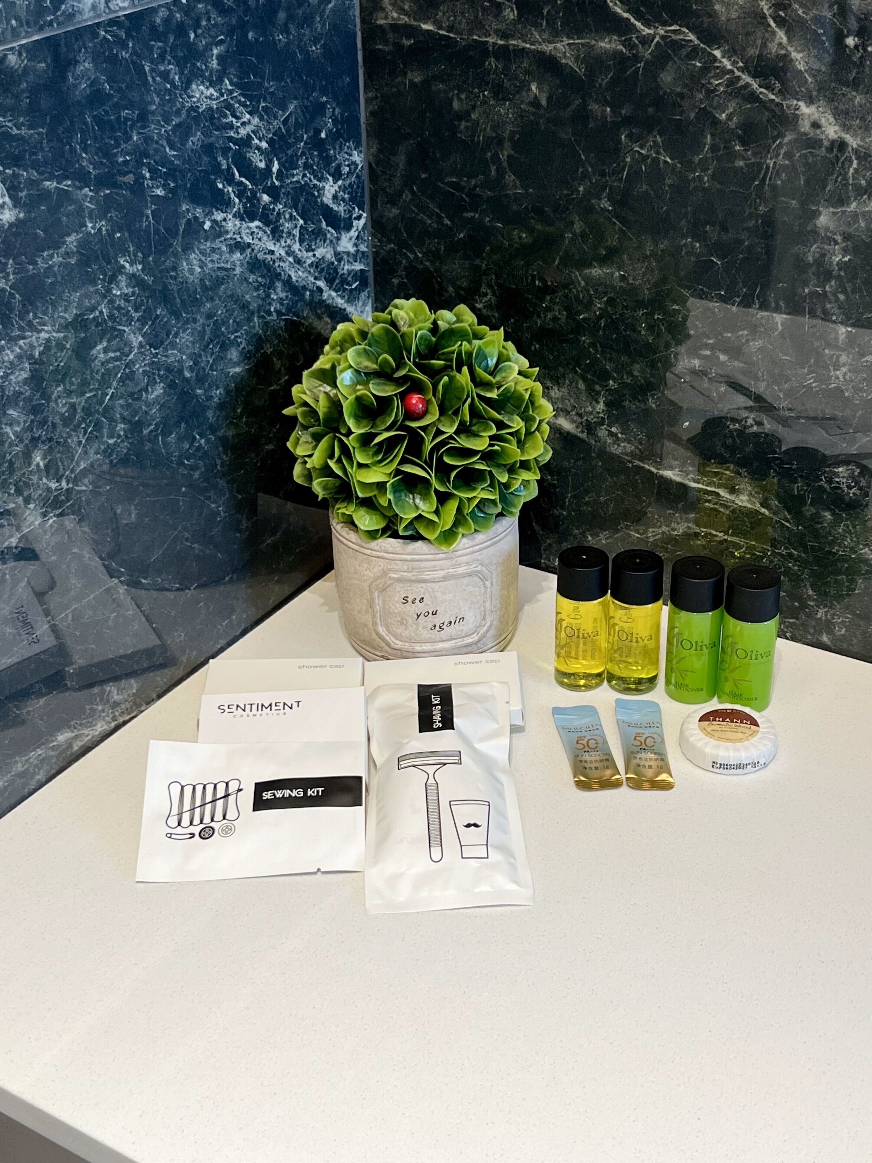 A bathroom vanity with a marble countertop, a potted plant, and toiletries including a shower cap, razor, shaving cream, and olive oil bottles.