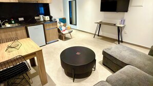 Una sala moderna con sofá, una mesa de centro, una mesa de comedor con sillas, una zona de cocina con refrigerador y un televisor de pantalla plana montado en la pared.