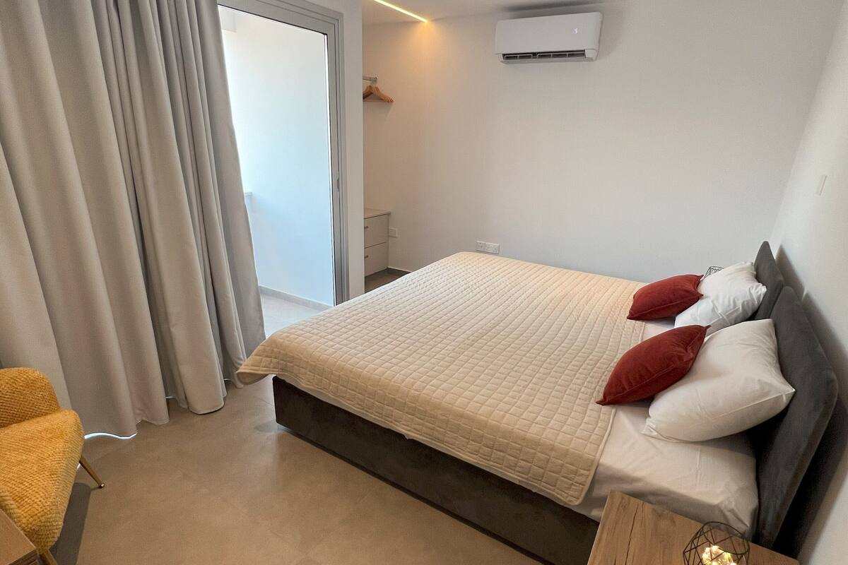 Een moderne slaapkamer met een groot bed, een houten nachtkastje, een stoel en uitzicht op de natuur via een schuifdeur.