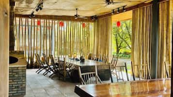 Un comedor con una mesa larga y sillas, una chimenea de piedra y vistas a través de cortinas de bambú.