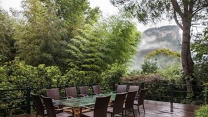 A wooden deck with a glass-top table and wicker chairs, surrounded by lush greenery and a view of a cliff. - Wuyishan Guanjing Shanfang Hotel (Wuyishan)