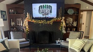 Un salon chaleureux avec une cheminée, un téléviseur diffusant un programme sur les stratégies offensives de l’équipe de l’Ohio, et une table basse sur laquelle se trouve un plateau avec divers objets.