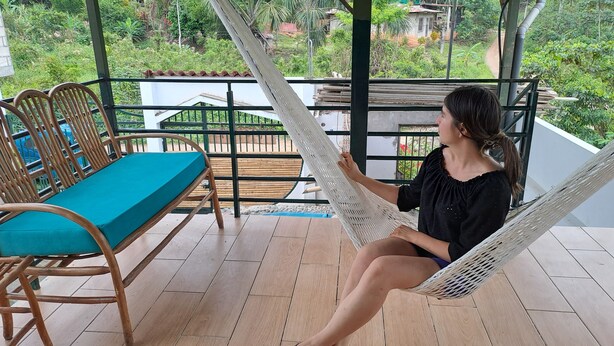 Un fauteuil suspendu avec un coussin turquoise, placé sur une terrasse en bois, d’où l’on peut admirer un jardin luxuriant et une maison au loin.