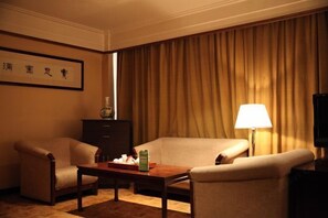 Una habitación de hotel con sofá, dos sillones, una mesita con una bandeja de aperitivos, una lámpara y un televisor.
