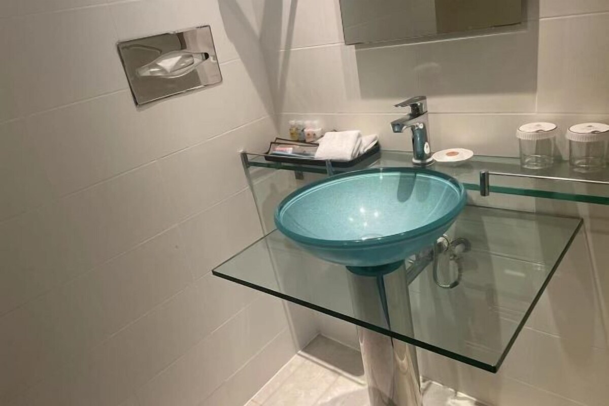 Une salle de bain moderne avec un lavabo en verre, un distributeur de serviettes et une étagère en verre où sont rangés les articles de toilette.