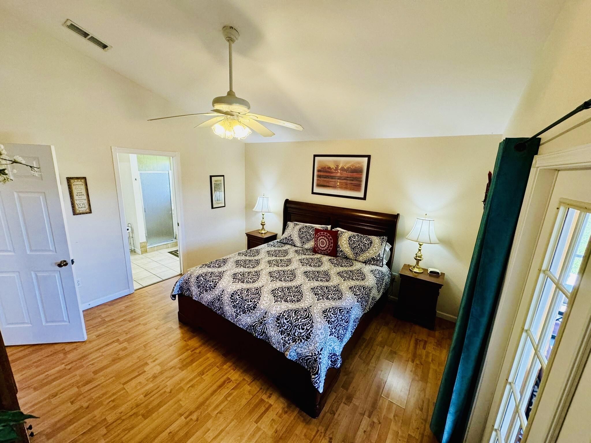 A bedroom with a wooden floor, a bed with patterned bedding, a ceiling fan, and a door leading to another room.