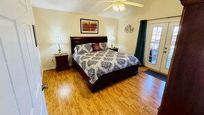 Une chambre avec un plancher en bois, un lit recouvert d’une literie à motifs, un ventilateur de plafond et une porte avec un panneau de verre.