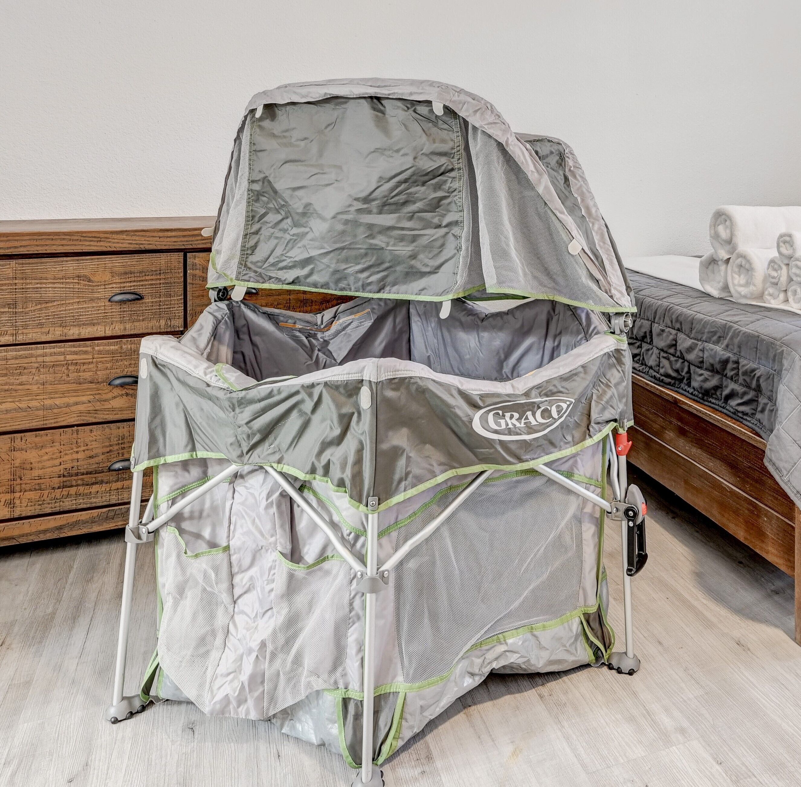 Een Graco babywieg met een kap, geplaatst in een kamer met een houten kastje en een bed met een gewatteerde spreidekker.