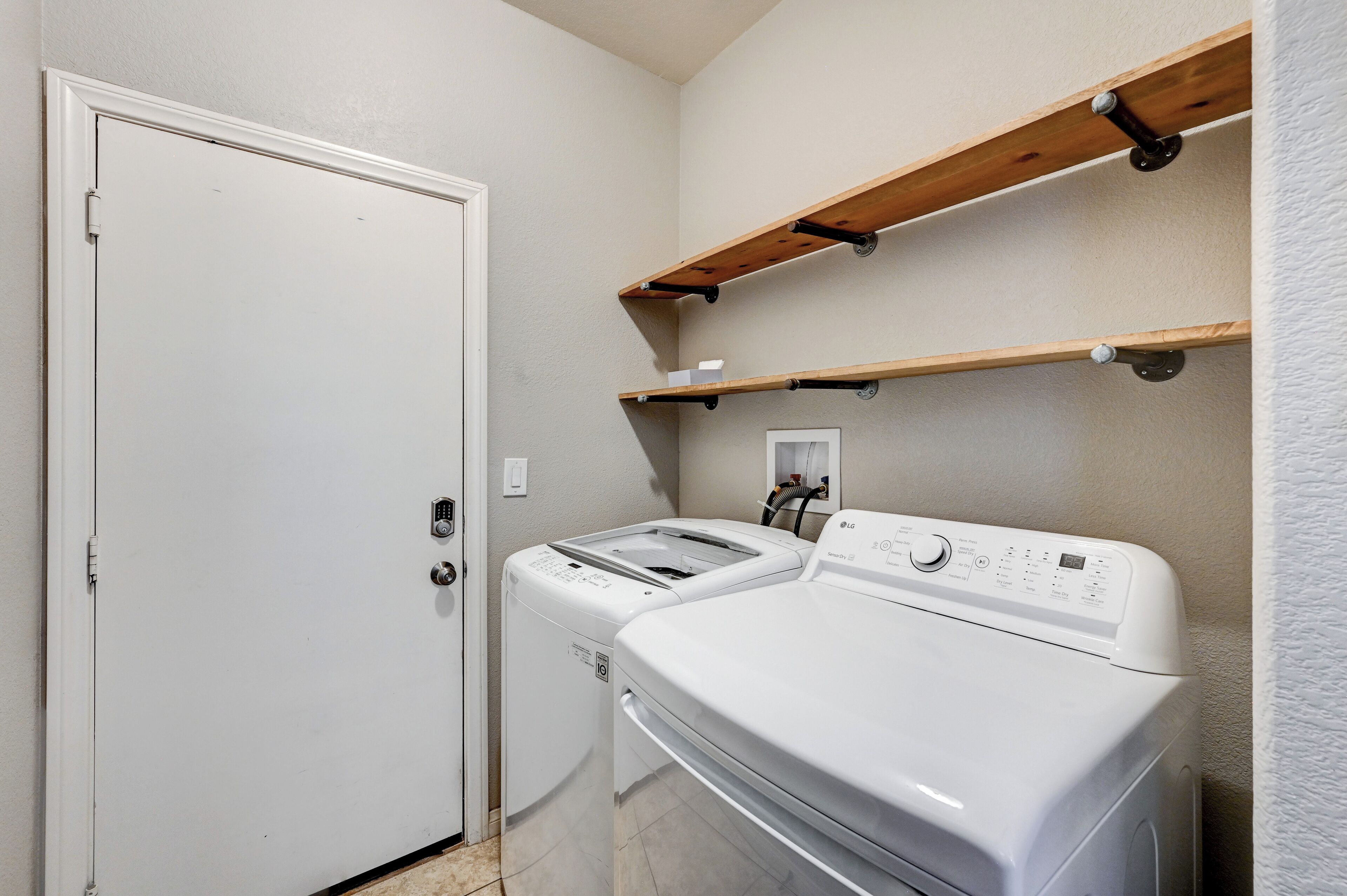 A laundry room with two white washing machines, wooden shelves above, and a door.