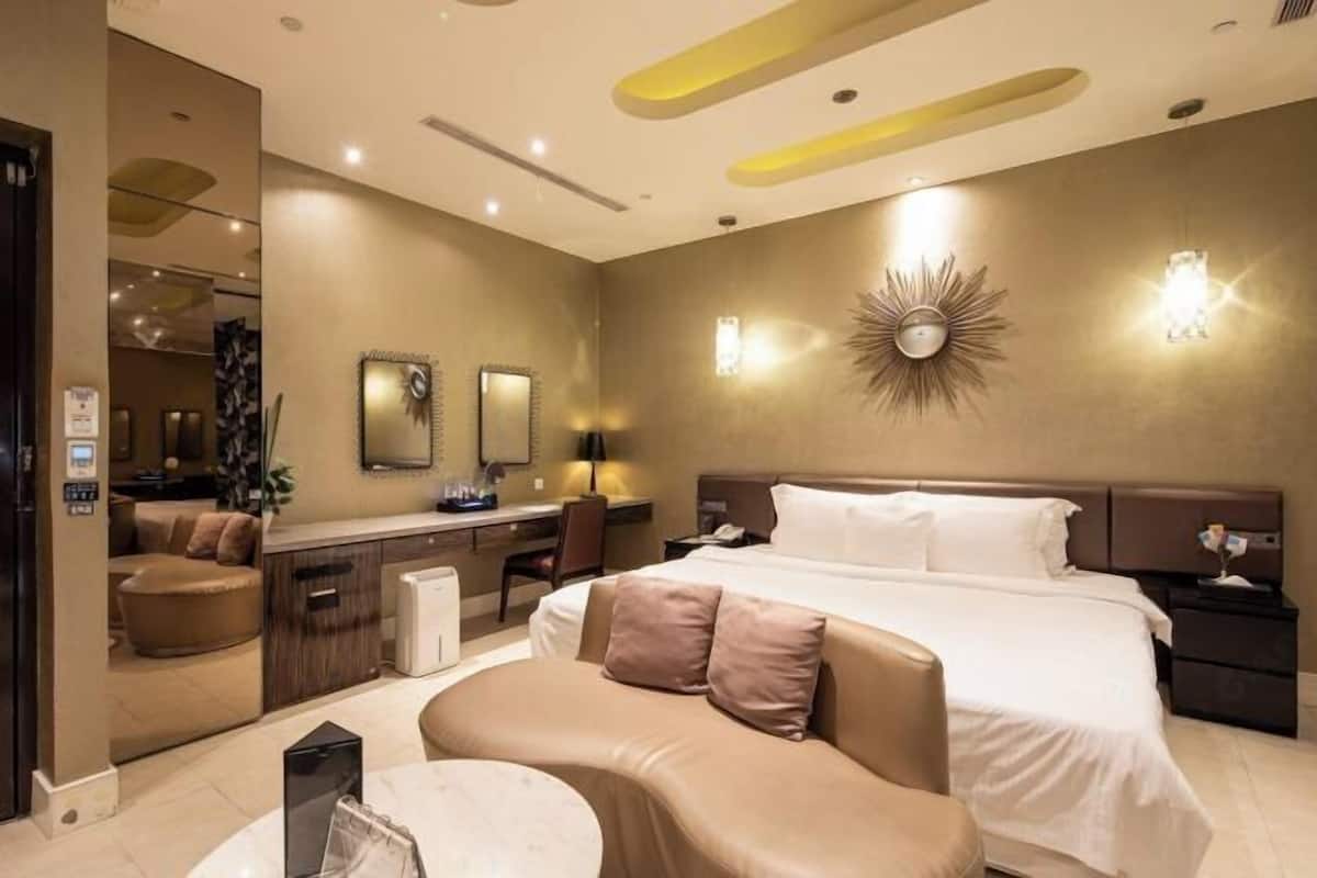 A modern hotel room with a large bed, a sitting area, a desk, and a mirror.