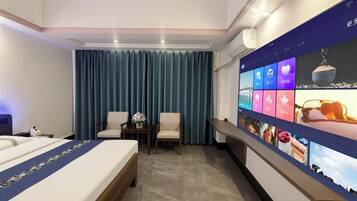 A hotel room with a bed, two chairs, a small table with a vase, a large digital screen displaying images, and a curtain.