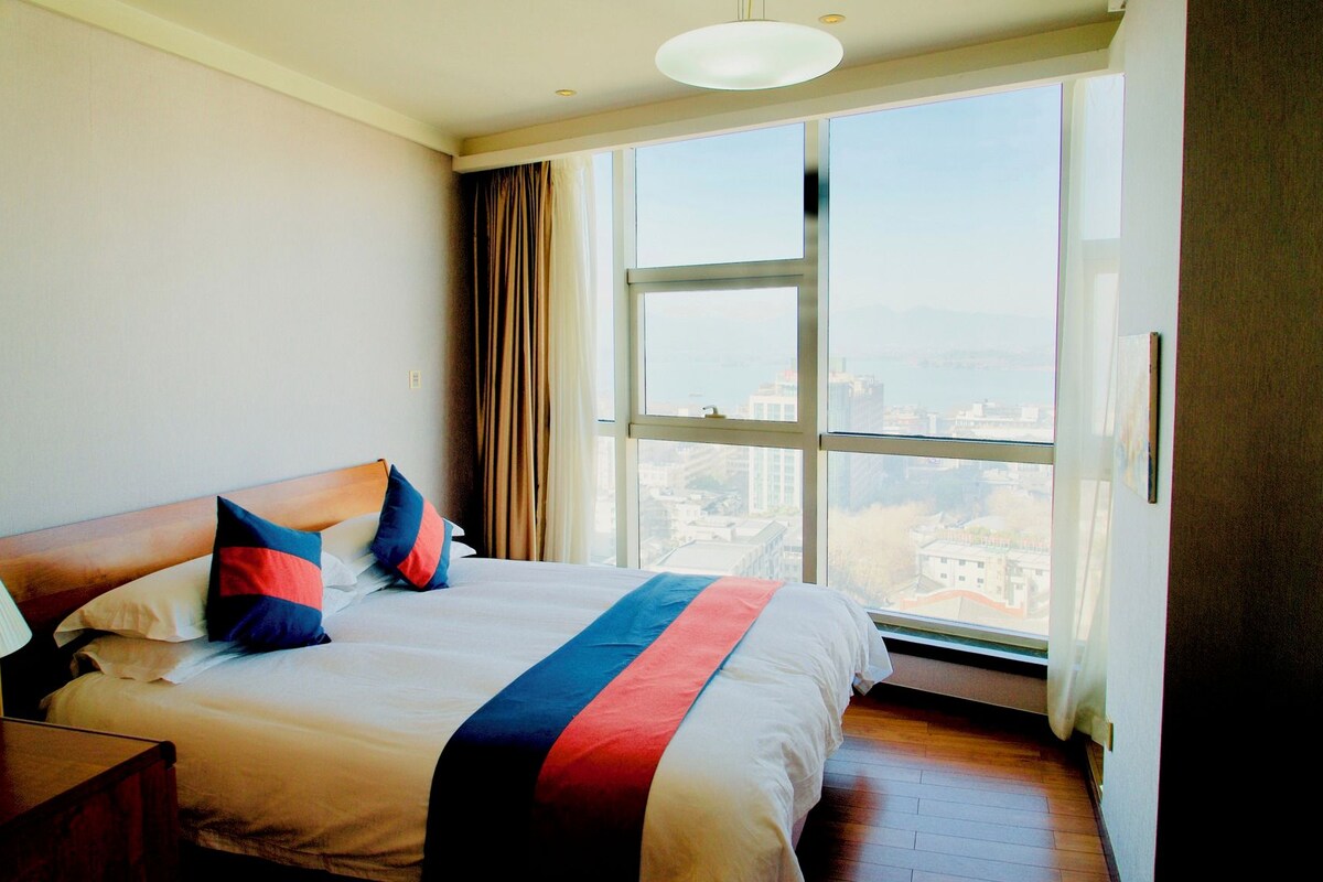 A hotel room with a large bed, a wooden headboard, and a view of a cityscape through a large window.