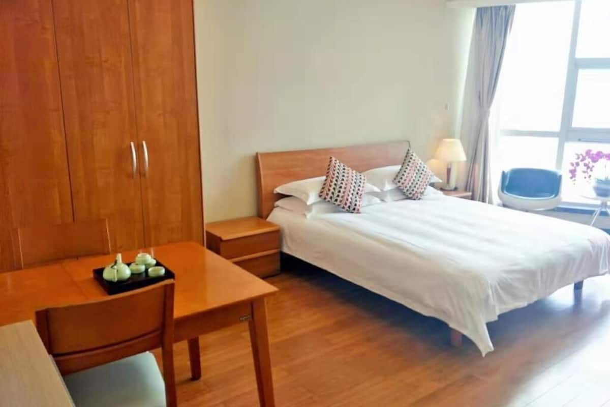 Una habitación de hotel con un armario de madera, una cama con ropa de cama blanca, una mesita de noche con una lámpara y vistas al exterior a través de una ventana.