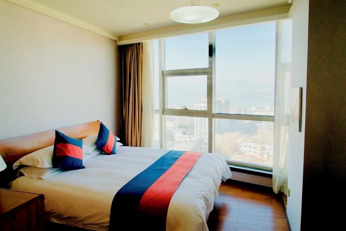 Habitación de hotel con una cama grande, una mesita de noche y vistas de la ciudad a través de un ventanal.