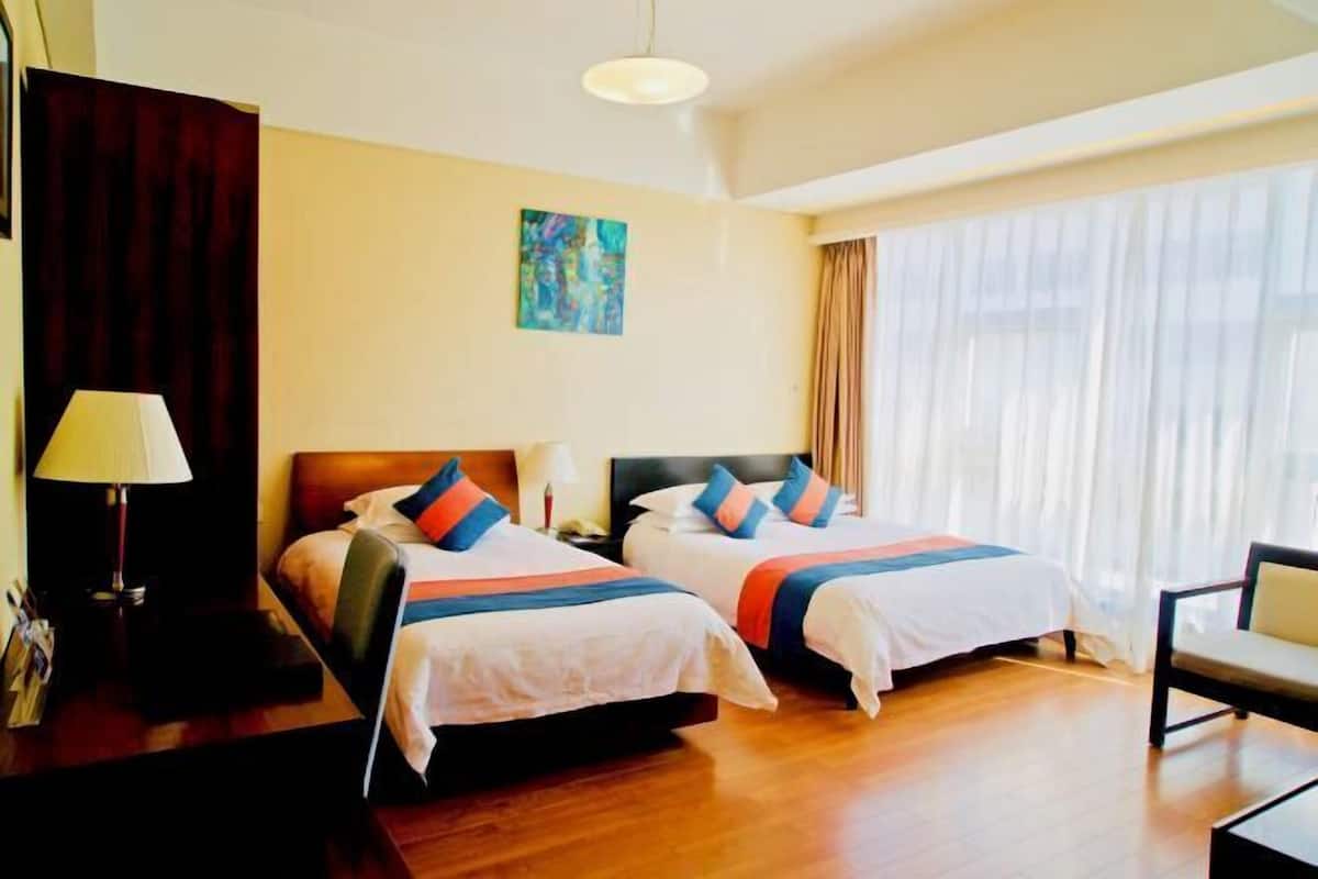 Habitación de hotel con dos camas, un escritorio con lámpara, una silla y un ventanal con cortinas.