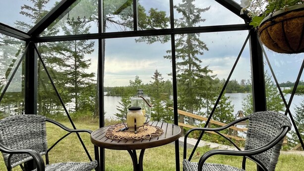 Un espace extérieur aménagé, abrité et doté de chaises et d’une table en osier, offrant une vue sur un lac et une forêt.