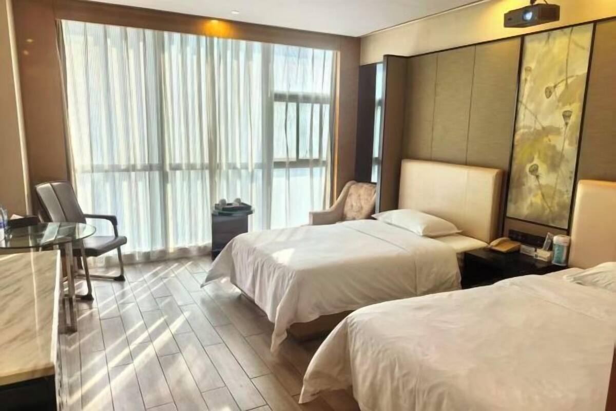 A hotel room with two beds, a desk, a chair, a large window with curtains, and a wall-mounted TV.