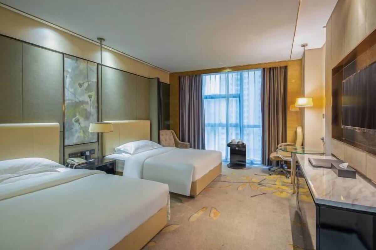 A hotel room with two beds, a desk, a chair, a TV, and a large window with curtains.