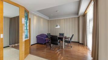 A hotel room with a wooden floor, a dining area with a table and chairs, a purple sofa, and a large window with curtains.