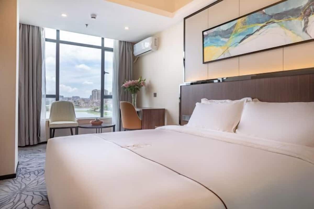 A hotel room with a large bed, a desk, a chair, and a view of the city through large windows.