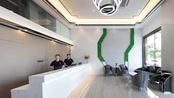 A modern reception area with a white reception desk, two staff members, a green decorative wall, and seating arrangements.