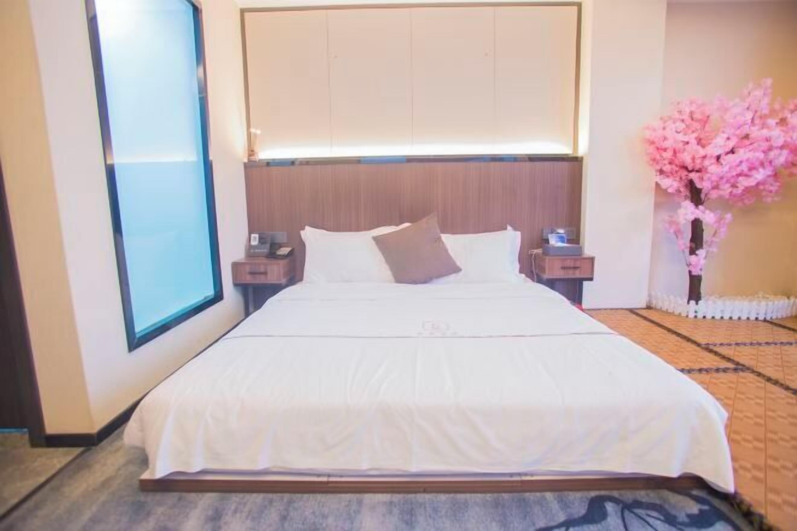 Habitación de hotel con una cama grande, una mesita de noche y una ventana, además de un árbol decorativo rosa.