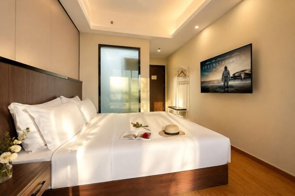 A hotel room with a large bed, a bedside table, a nightstand, a wall-mounted TV, and a door leading to another room.