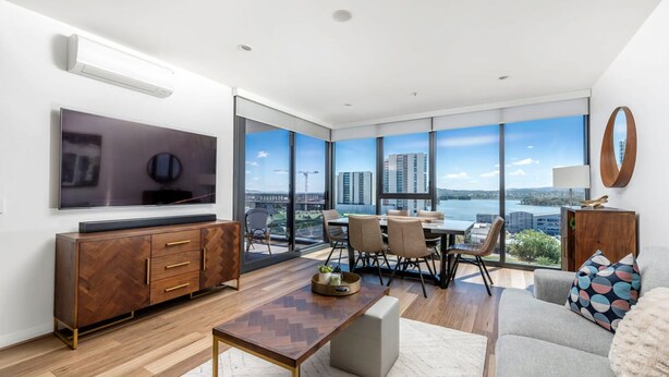 Una sala de estar moderna con un televisor grande, muebles de madera, un comedor y vistas a un paisaje urbano a través de amplias ventanas.