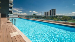 Una piscina azul cristalina con cubierta de madera y barandillas de vidrio, con vistas a un paisaje urbano con edificios modernos.