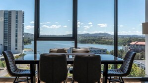 Comedor con una mesa y sillas, ventanas amplias que ofrecen vista a un lago y edificios.