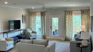 A spacious living room with a flat-screen TV, grey sofas, a coffee table, and a wicker ottoman.