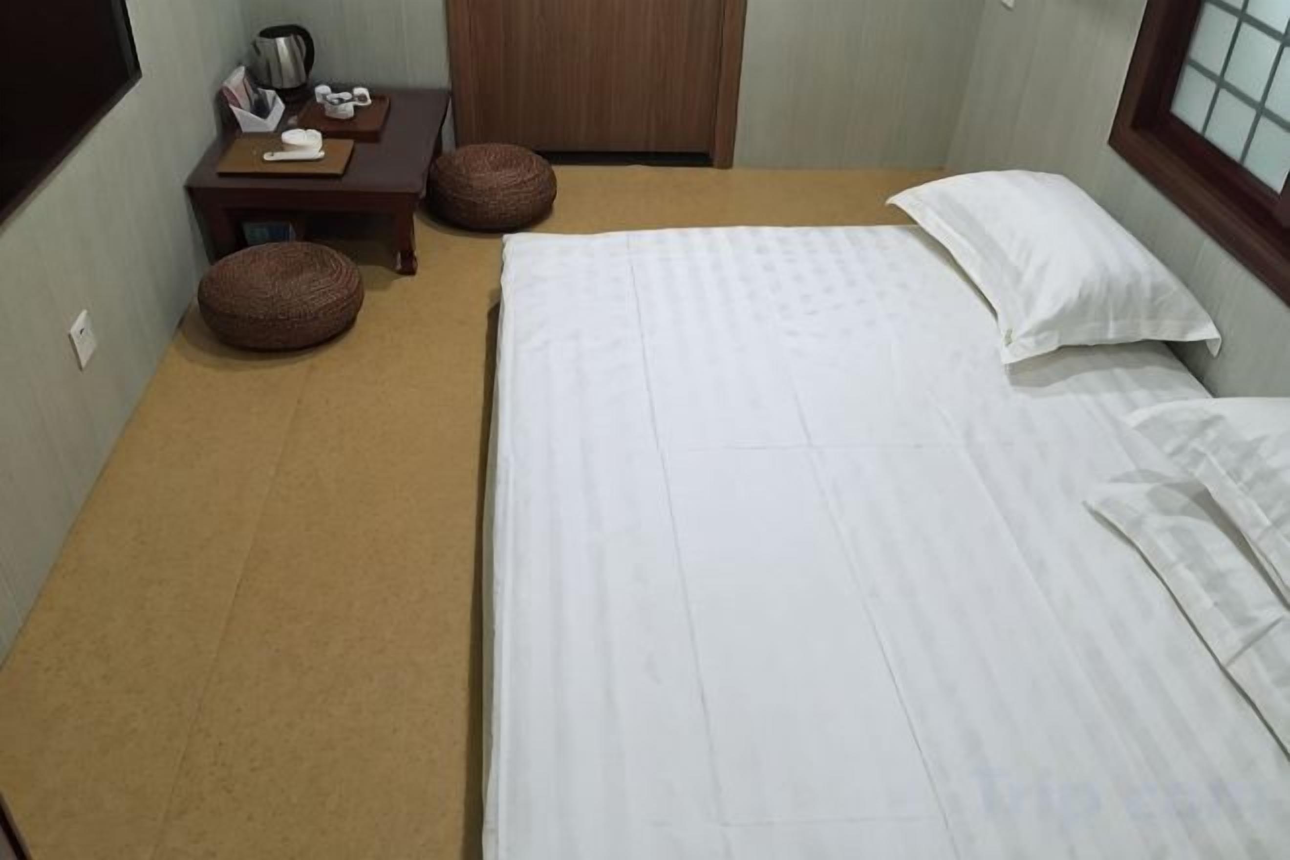 A neatly made bed with white linens, a small wooden nightstand with a lamp, and a brown ottoman.