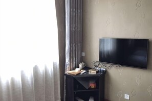 Una habitación con una televisión montada en la pared, una mesita con un libro y una taza, y una ventana con cortinas.