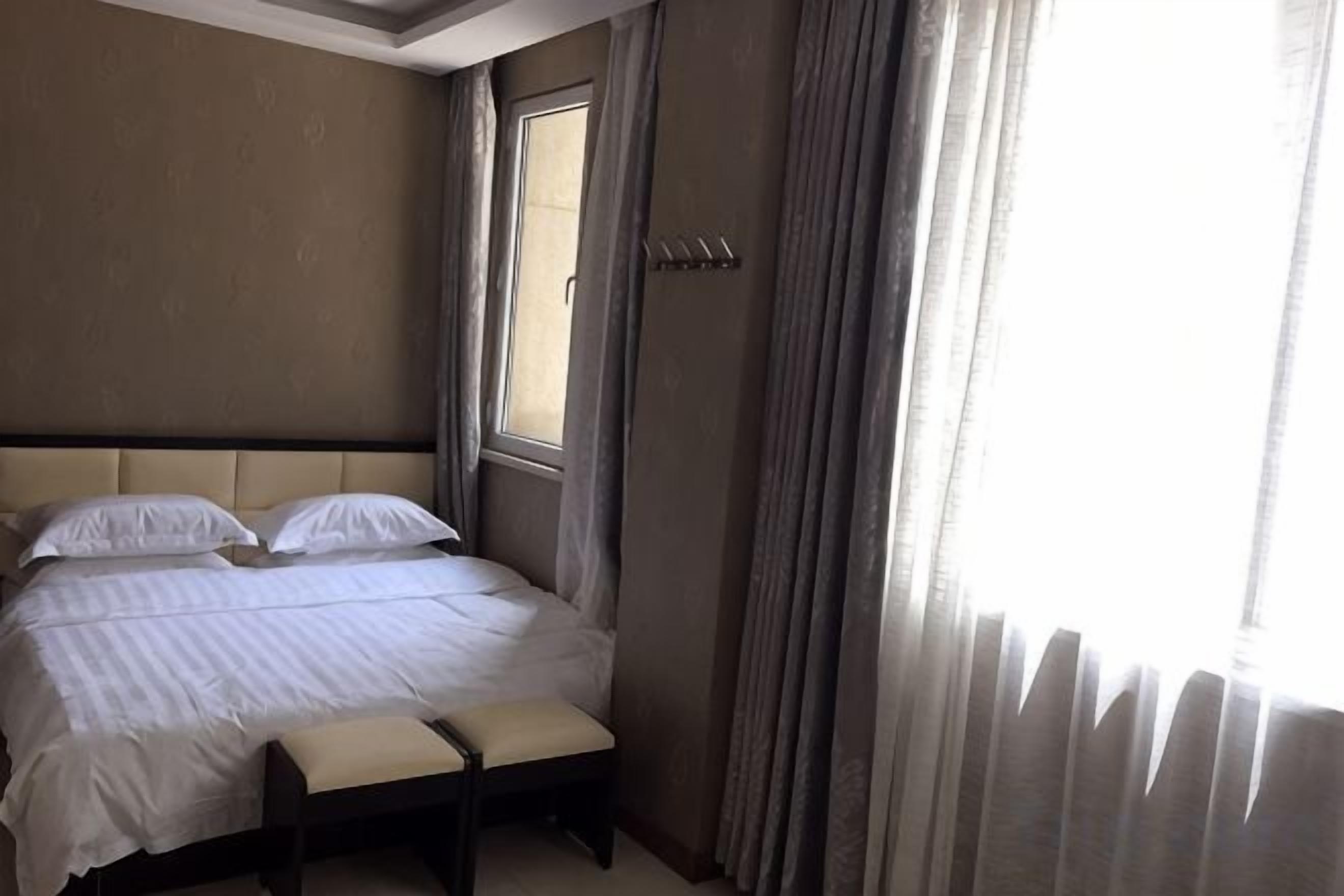 A hotel room with a bed, a bench, a window, and curtains.