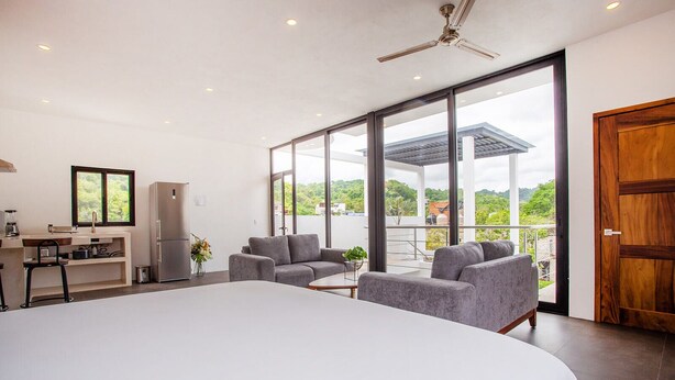Une chambre moderne avec un grand lit, des canapés gris et un balcon offrant une vue sur la verdure.