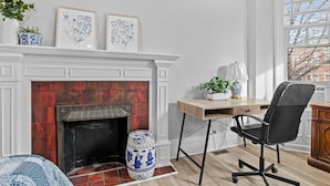 A room with a fireplace, a desk with a chair, a window with blinds, and a plant on the windowsill. - Beautiful Green-Door Brownstone (Jersey City)