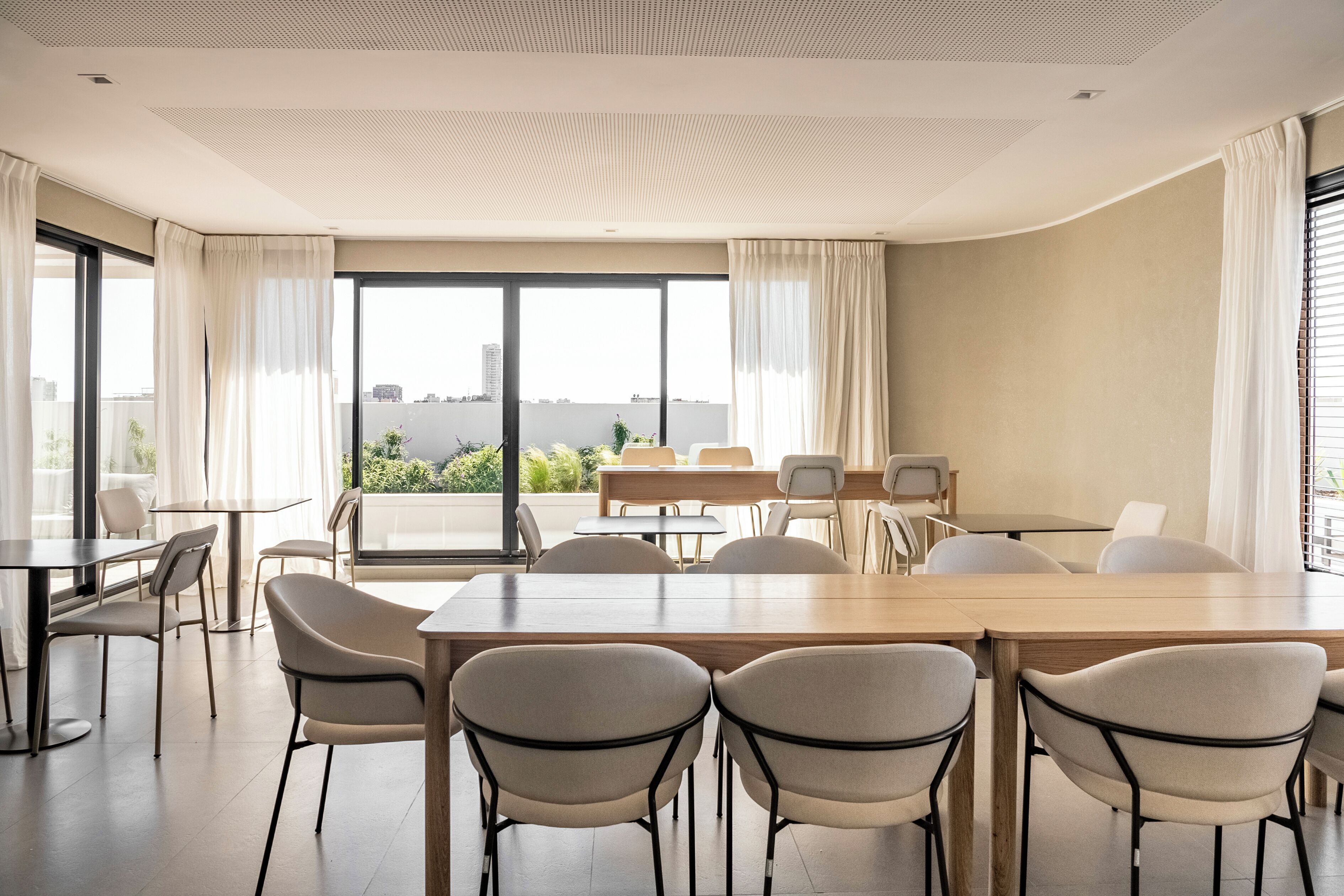 Uma sala de conferência moderna com uma grande mesa e cadeiras, grandes janelas com cortinas e vista para a paisagem urbana.