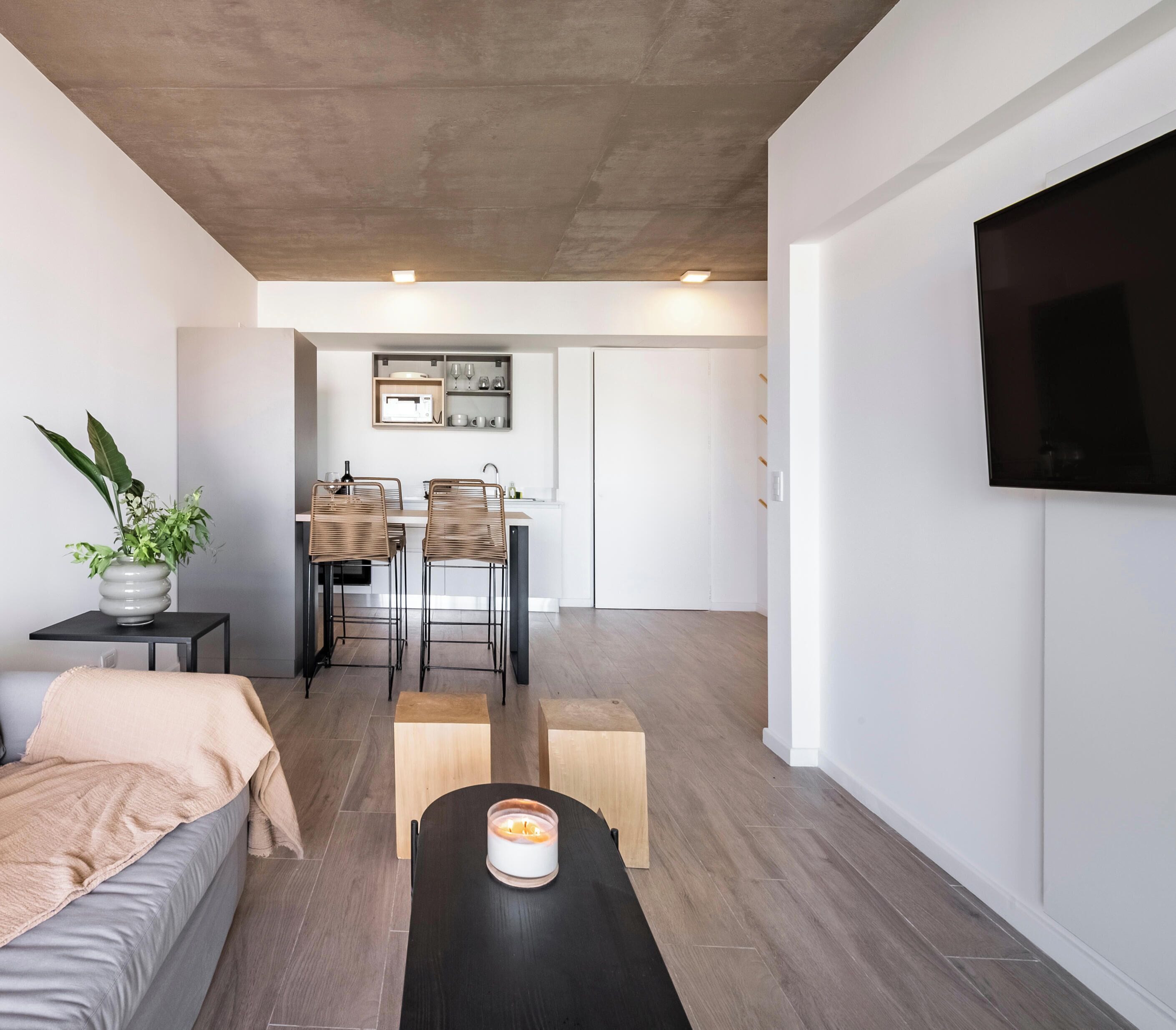 Un espacio de estar moderno con sofá, mesa de centro, comedor con sillas, cocina con microondas y televisor montado en la pared.