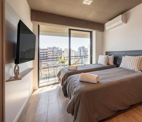 A hotel room with two beds, a TV on the wall, a balcony with a chair, and a city view. - Top Rentals Nuñez (Buenos Aires)